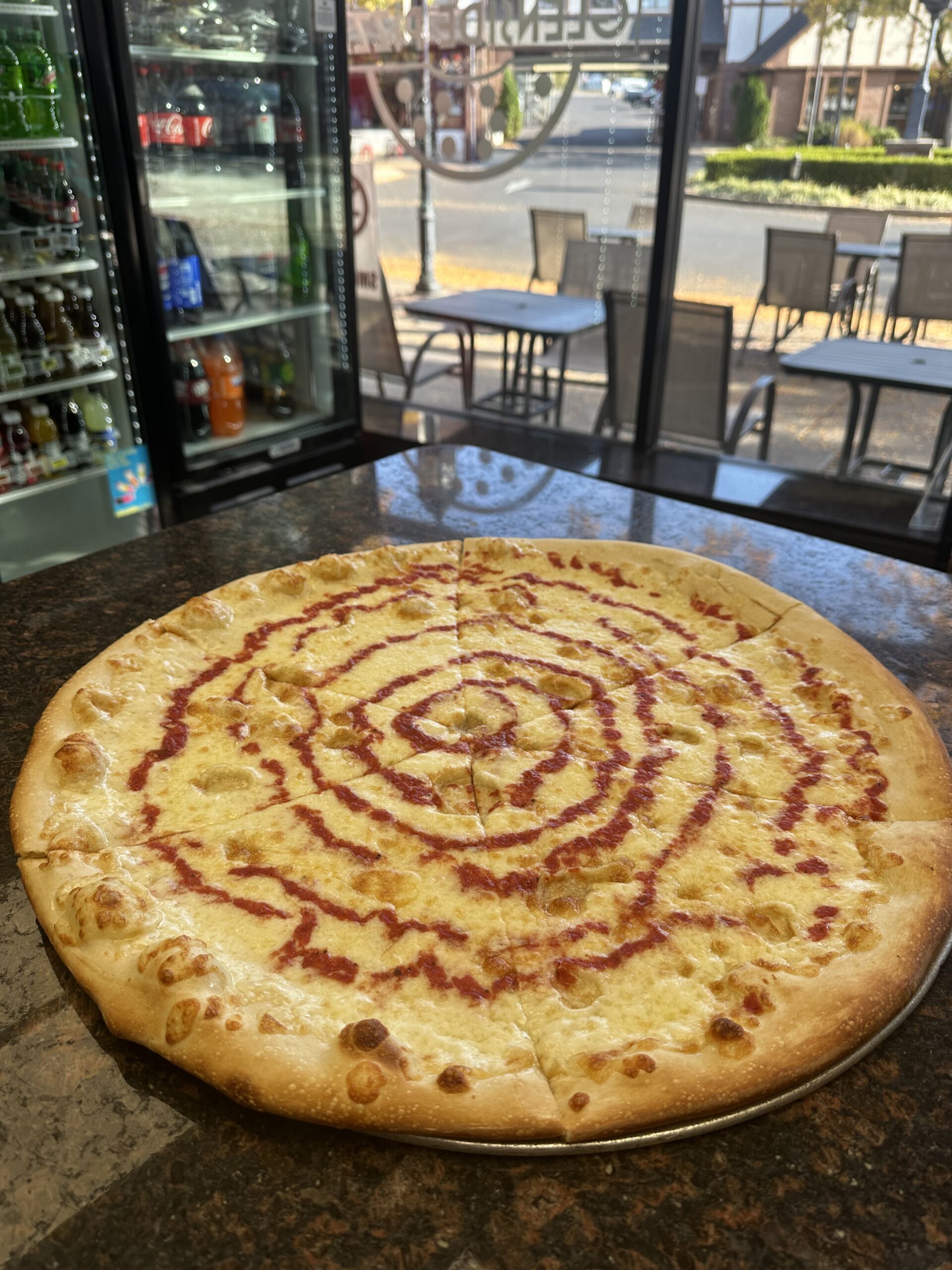 Large cheese pizza with spiral ketchup design on a counter, beverage fridge and outdoor seating visible in the background.