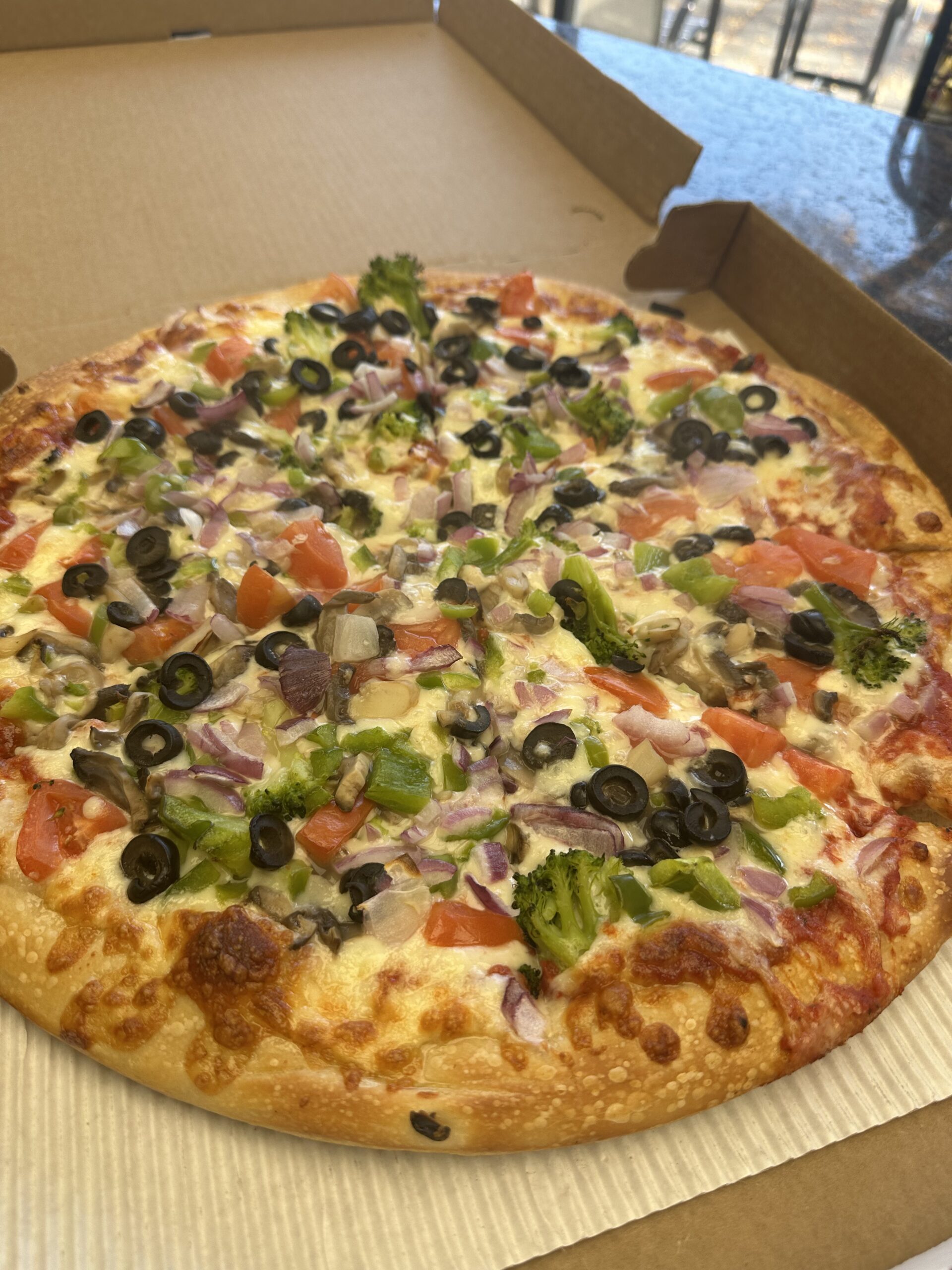 Vegetable pizza with olives, tomatoes, onions, broccoli, and green peppers in an open cardboard box.