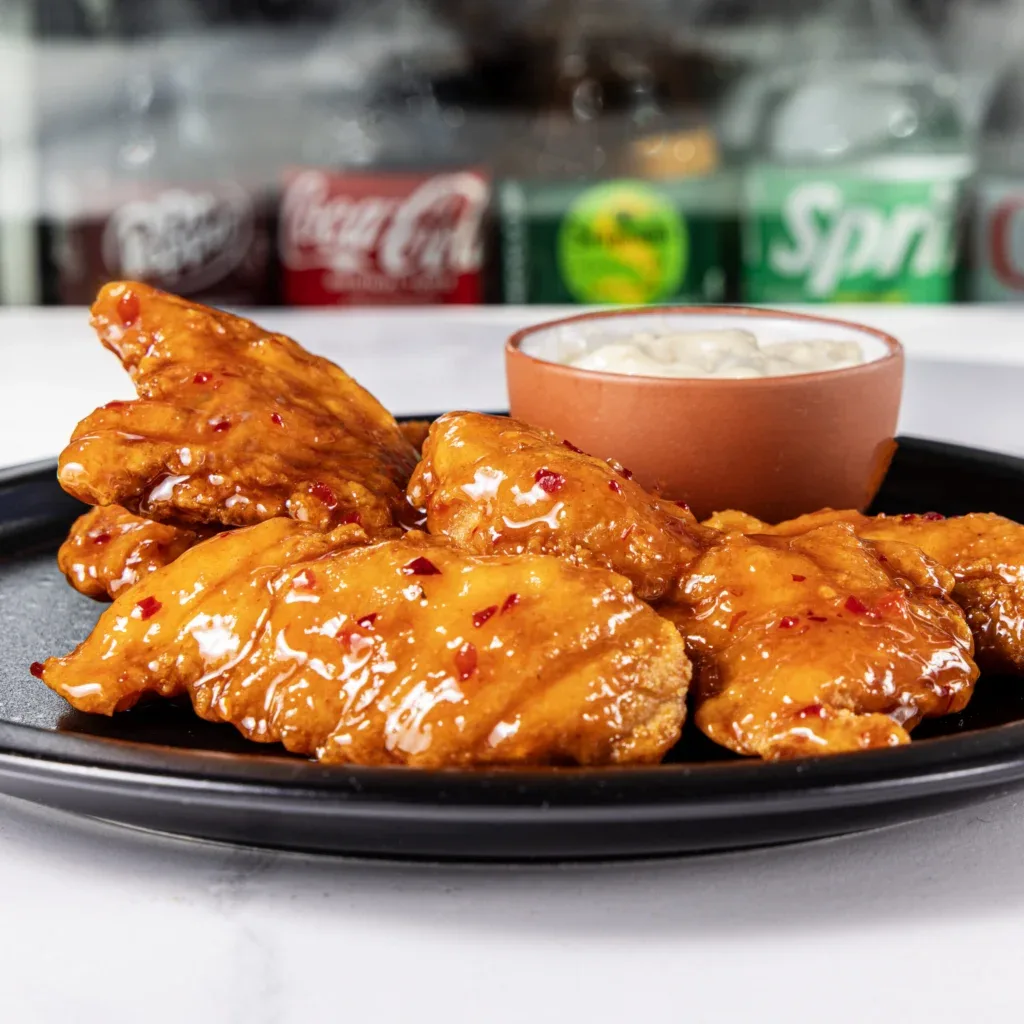 Glazed chicken wings with dipping sauce on a black plate, soda bottles in the background.