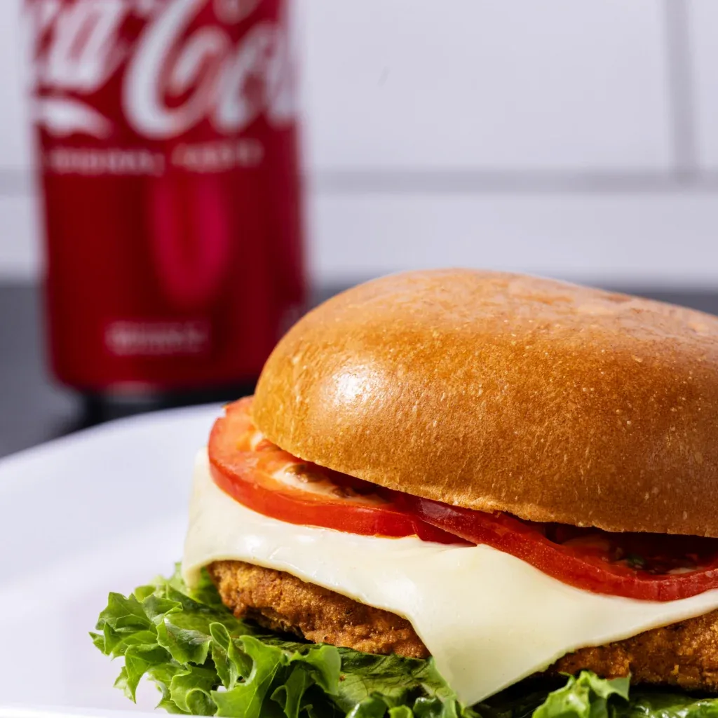 Chicken sandwich with lettuce, tomato, and cheese on a bun, with a Coca-Cola can in the background.